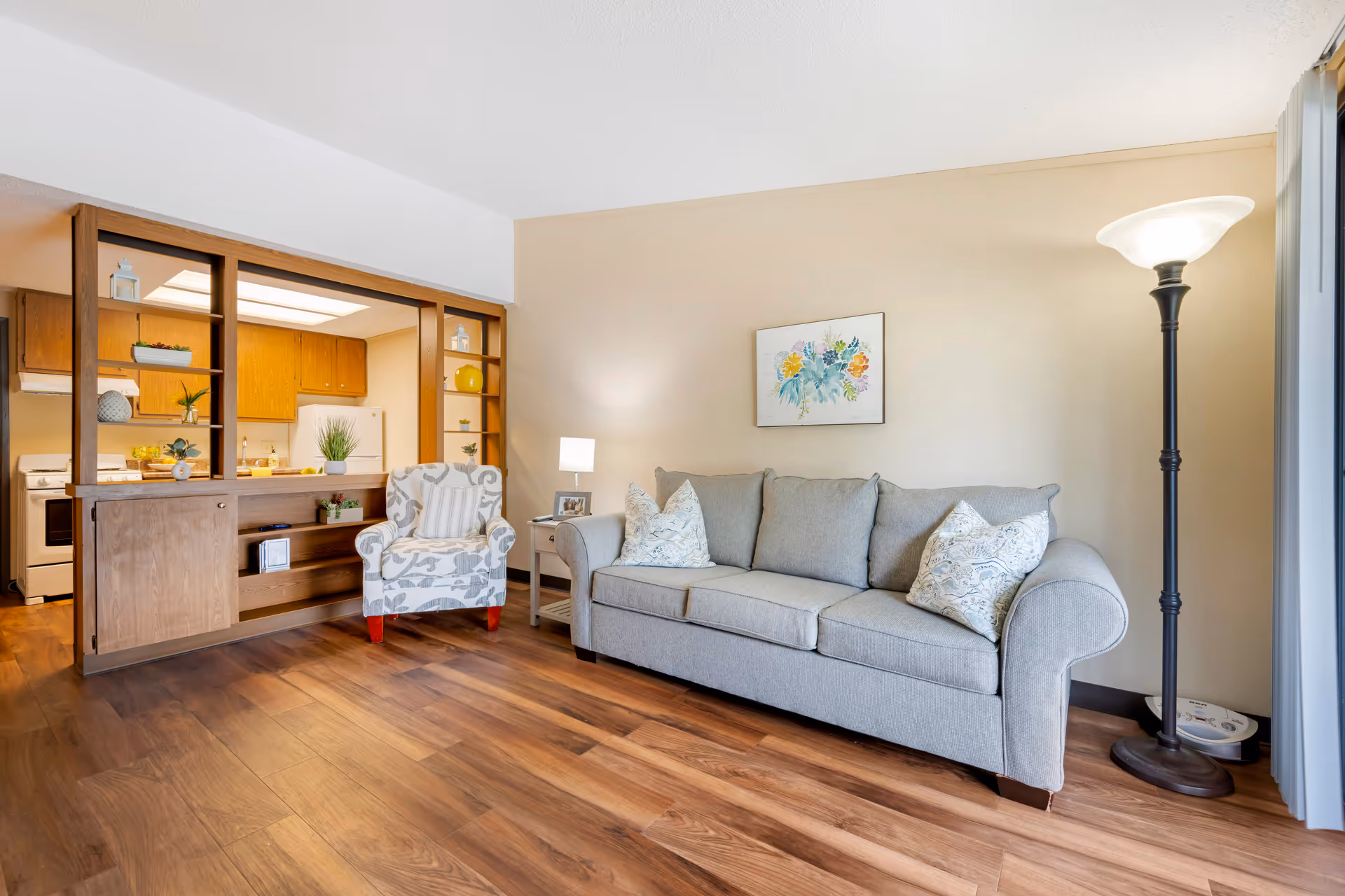 A cozy living room with a light gray sofa adorned with patterned pillows, a matching armchair, a small side table with a lamp and photo frame, and a tall floor lamp. The room has wooden flooring and beige walls with a floral painting. An open wooden shelving unit separates the living room from a kitchen area with wooden cabinets and white appliances.