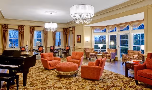 A spacious, well-lit living room area with large windows and glass doors letting in natural light. The room features multiple comfortable orange armchairs arranged around a round ottoman, a grand piano on the left side, and several small tables with chairs near the windows. Elegant chandeliers hang from the ceiling, and the room is decorated with patterned carpet and beige walls with molding details.