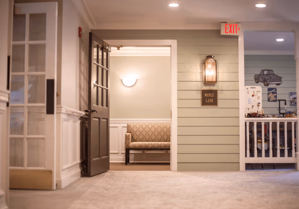 Interior hallway of a senior living facility with a partially open dark wooden door leading to a small seating area with a patterned bench. The hallway has light green walls with white wainscoting and a wall-mounted lantern light above a sign that reads 'MAPLE LANE'. To the right, there is a railing and a wall decorated with vintage-style signs and a truck illustration. An illuminated exit sign is visible above the hallway.