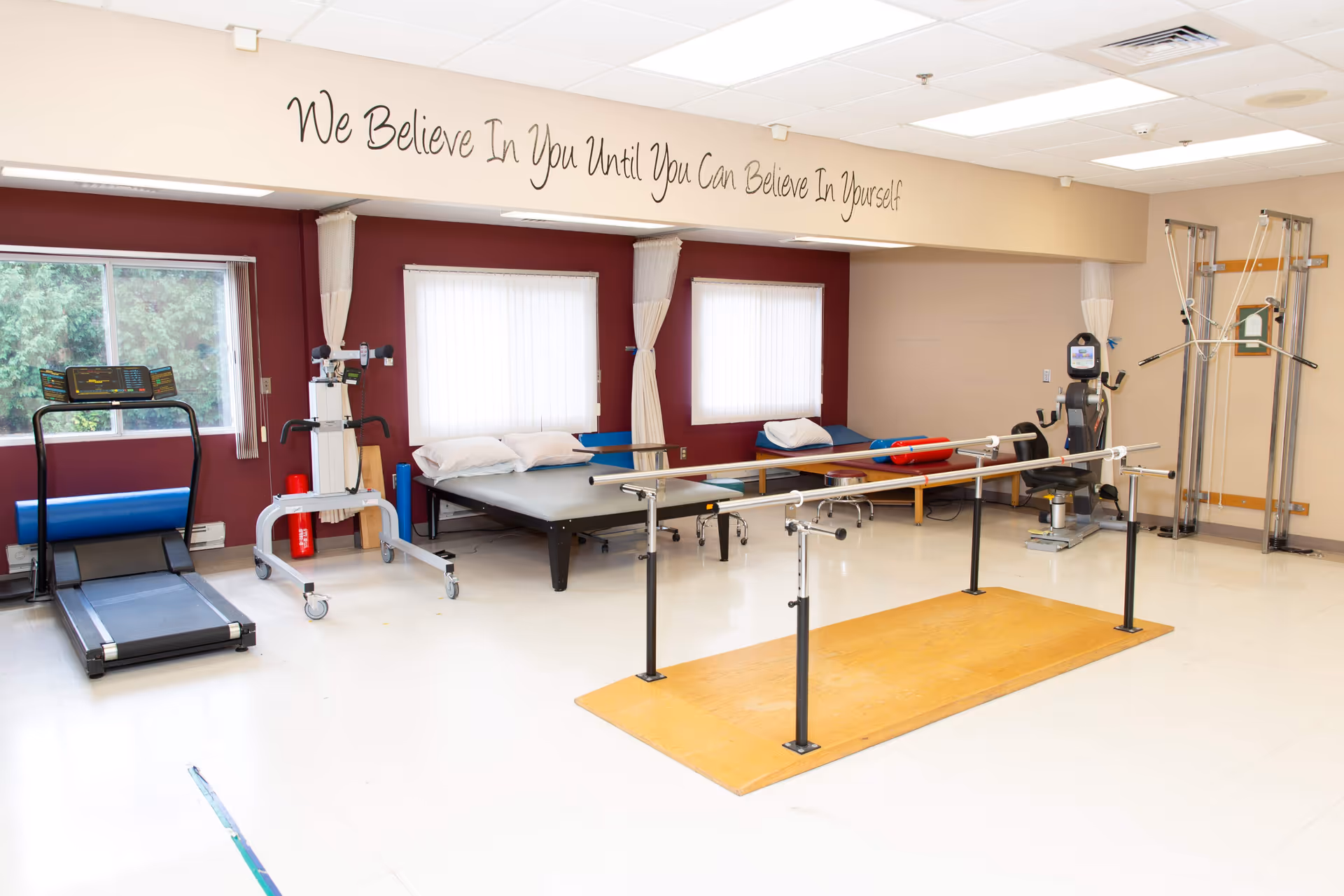 Rehabilitation room with treadmill, parallel bars, therapy tables and various exercise equipment under a wall quote.