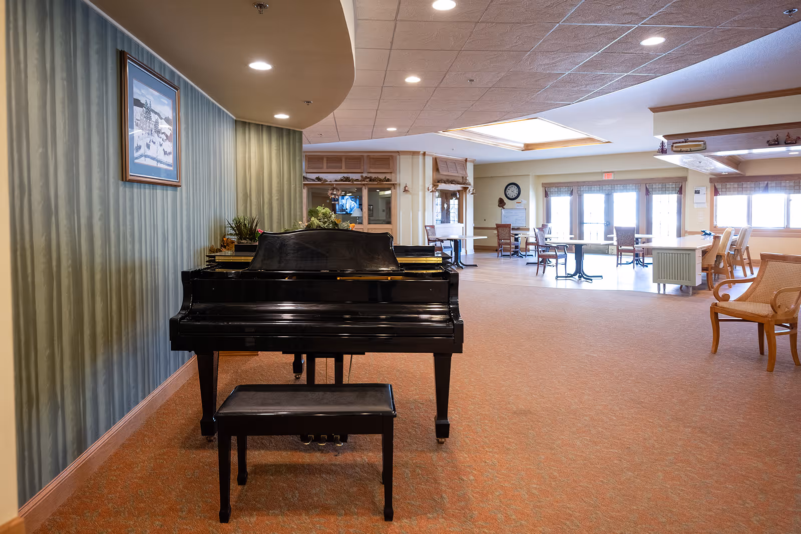 Interior view of a senior living facility common area featuring a black grand piano with a matching bench in the foreground. The room has carpeted floors, greenish striped wallpaper on one wall, and recessed ceiling lights. In the background, there are several tables and chairs near large windows and glass doors letting in natural light.