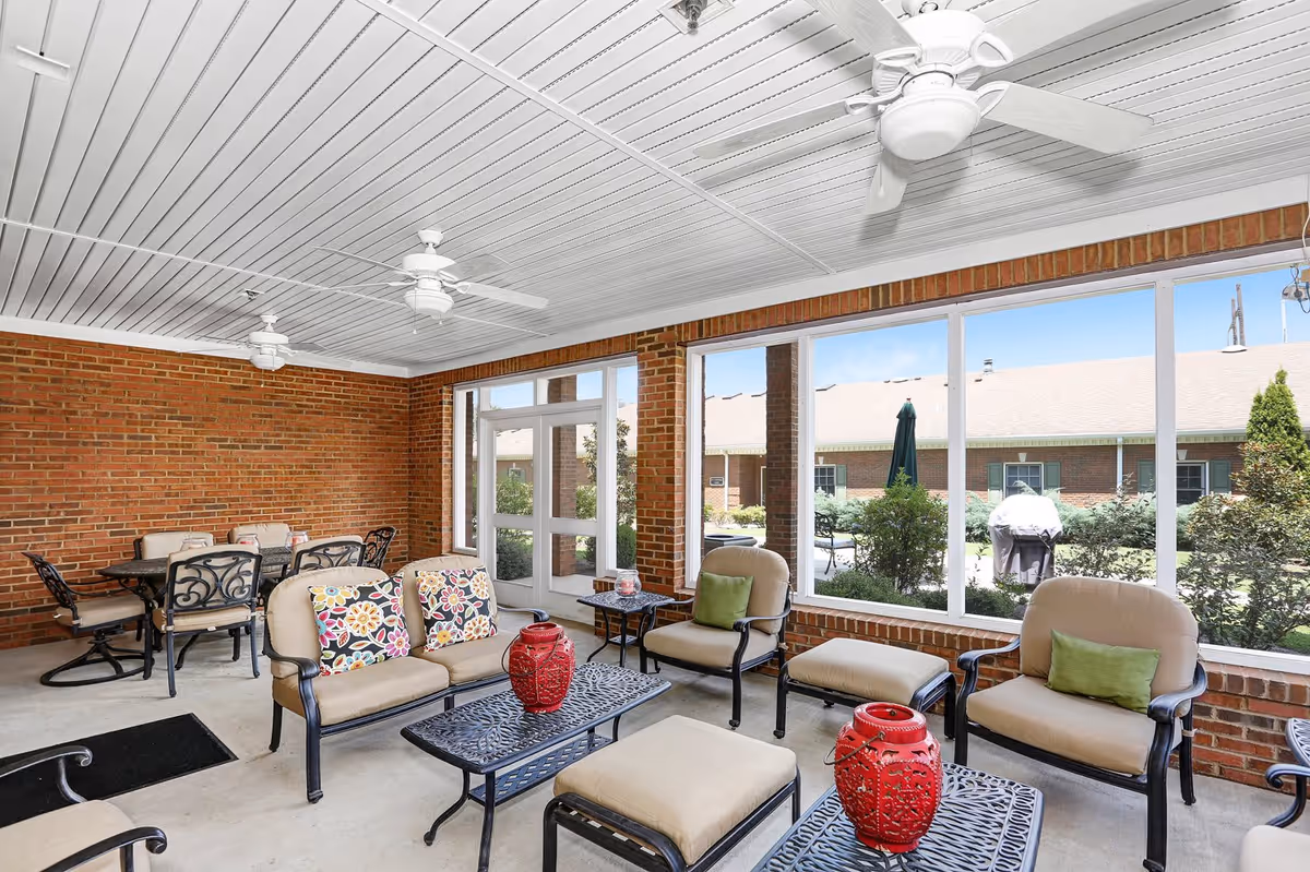 A bright and spacious indoor seating area with brick walls and large windows overlooking an outdoor garden. The room features cushioned chairs and sofas with colorful pillows, black metal tables with red decorative lanterns, ceiling fans, and a dining table with chairs in the corner.