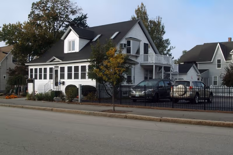 A two-story white house with a black roof, featuring a small balcony and several windows. There are two cars parked in a fenced parking area beside the house. Trees and shrubs surround the property, and the scene is set on a quiet street with neighboring houses visible in the background.