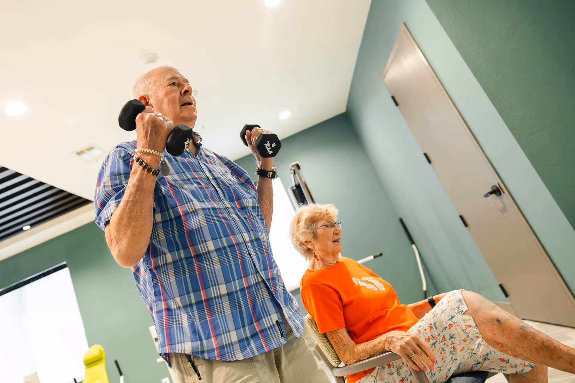An elderly man lifting dumbbells while an elderly woman sits nearby in a chair, both in a well-lit room with green walls and a closed door in the background.