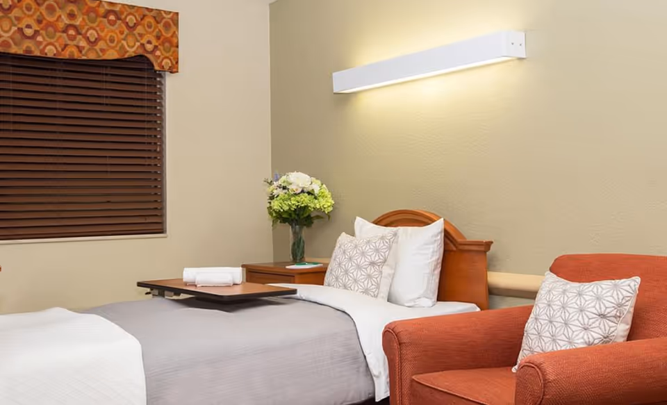 Senior living facility bedroom featuring a single bed, nightstand with flowers, and a red chair.