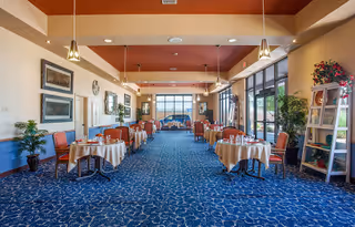 A spacious dining room with multiple round tables covered with beige tablecloths and set with plates and cups. The room has large windows on the right side letting in natural light, blue patterned carpet, and warm beige walls with framed artwork. There are pendant lights hanging from the ceiling and some plants and decorative items placed around the room.