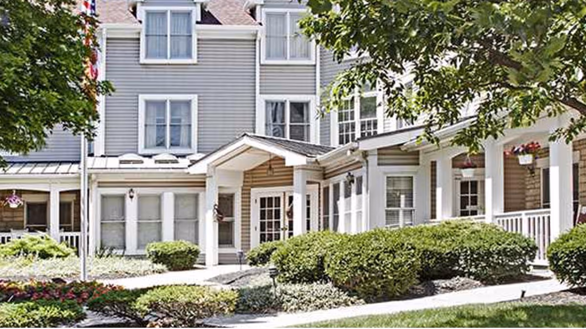 Exterior view of a senior living facility building with gray siding, white trim, and multiple windows. The entrance has a covered porch with white columns, surrounded by well-maintained bushes, trees, and landscaped flower beds.