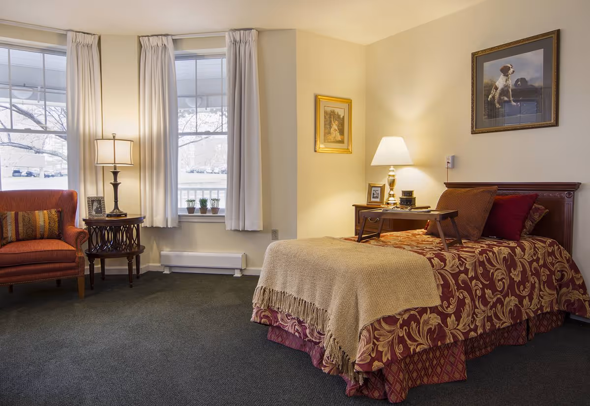 A cozy bedroom in a senior living facility featuring a single bed with a patterned bedspread and beige throw blanket. A wooden tray with a lamp and small items sits on the bed. The room has two large windows with white curtains, a red upholstered armchair with a decorative pillow, a round wooden side table with a lamp and framed photo, and framed artwork on the walls.