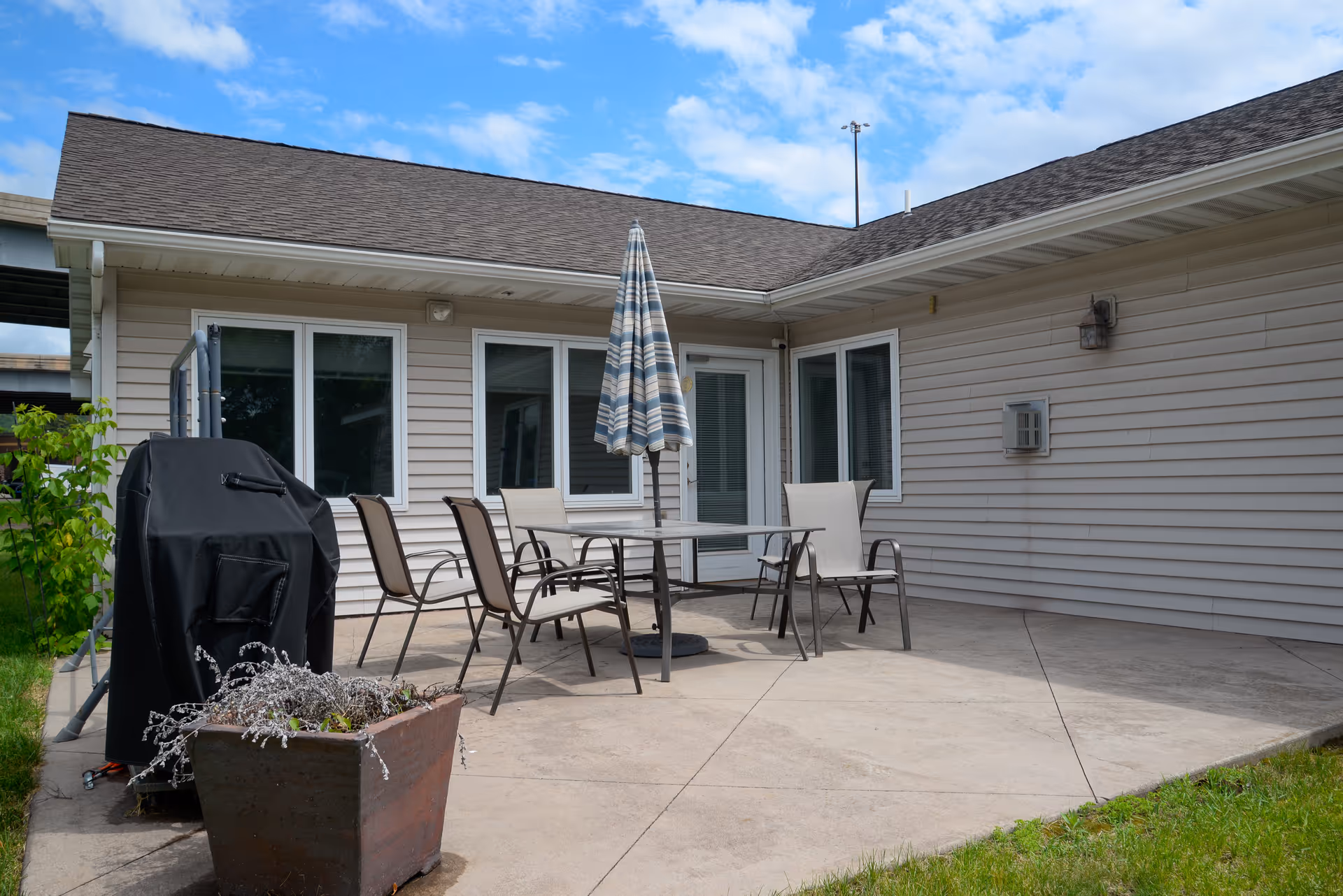 Outdoor patio area with a table, six chairs, and a closed striped umbrella. There is a covered grill on the left side and a large planter with some plants in front. The patio is adjacent to a beige siding building with several windows and a door, under a partly cloudy blue sky.