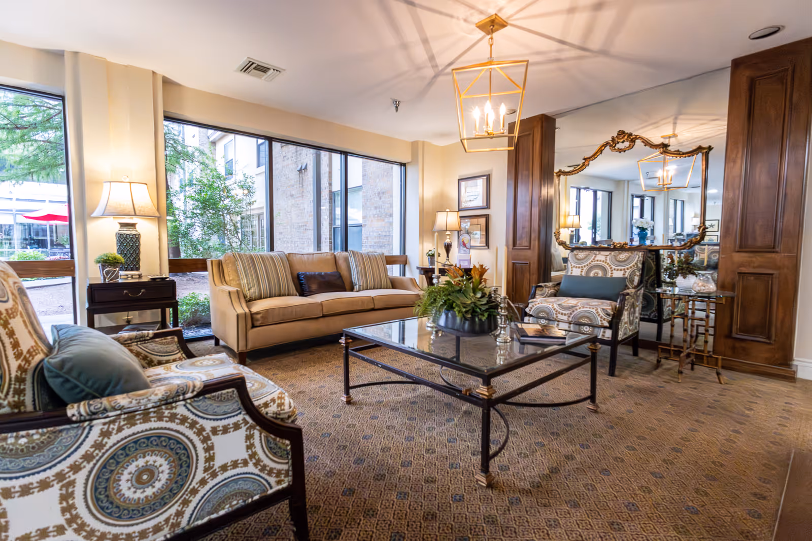 A cozy living room area with a beige sofa, two patterned armchairs with blue cushions, a glass coffee table with a plant centerpiece, and a large ornate mirror on the wall. The room has large windows letting in natural light, a carpeted floor, and warm lighting from table lamps and a ceiling fixture.