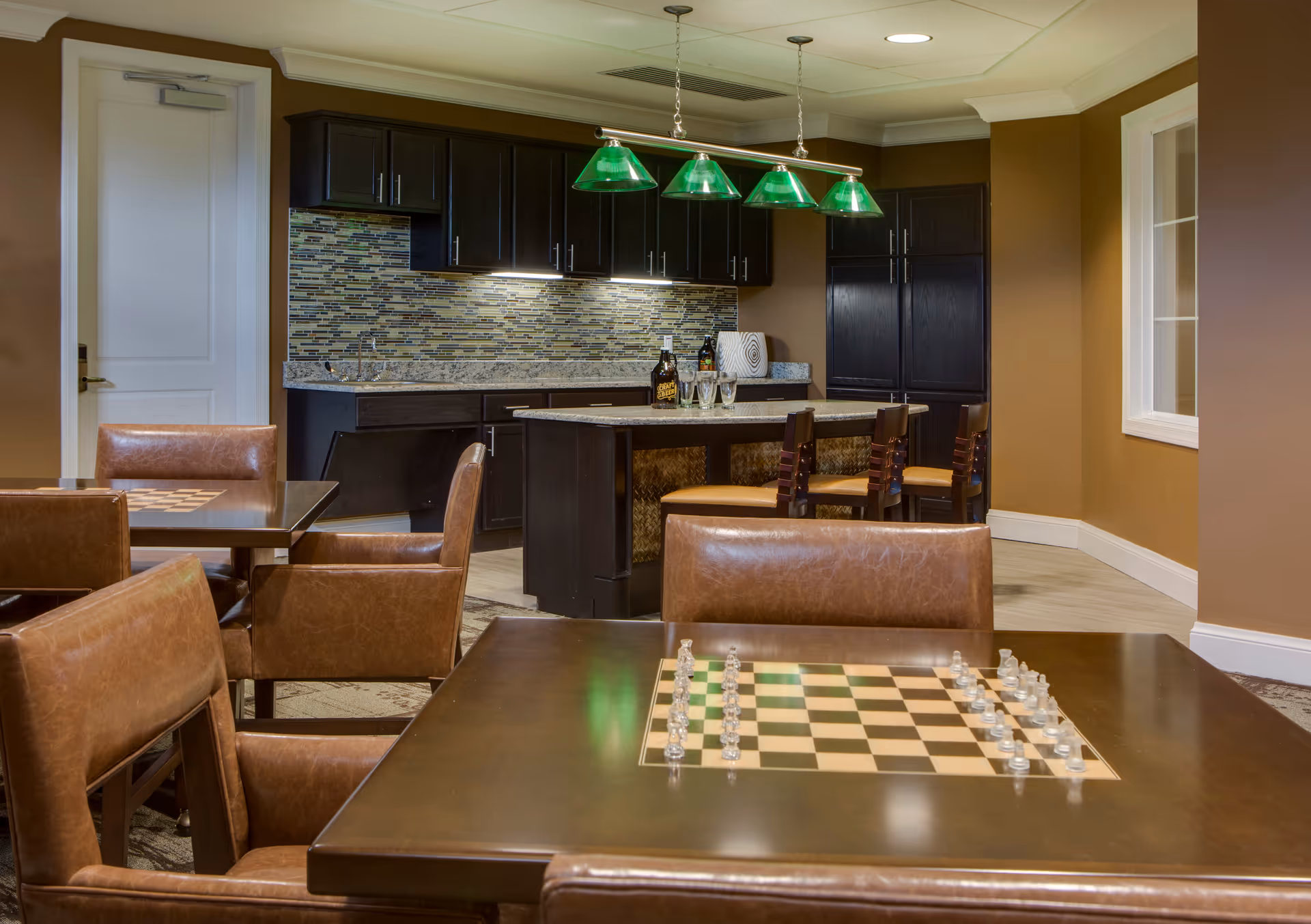 Interior view of a common area with a kitchen and seating. The kitchen features dark cabinets, a mosaic tile backsplash, and a central island with three chairs. In the foreground, there are tables with built-in chess boards and brown leather chairs. The room has warm brown walls and a window on the right side.
