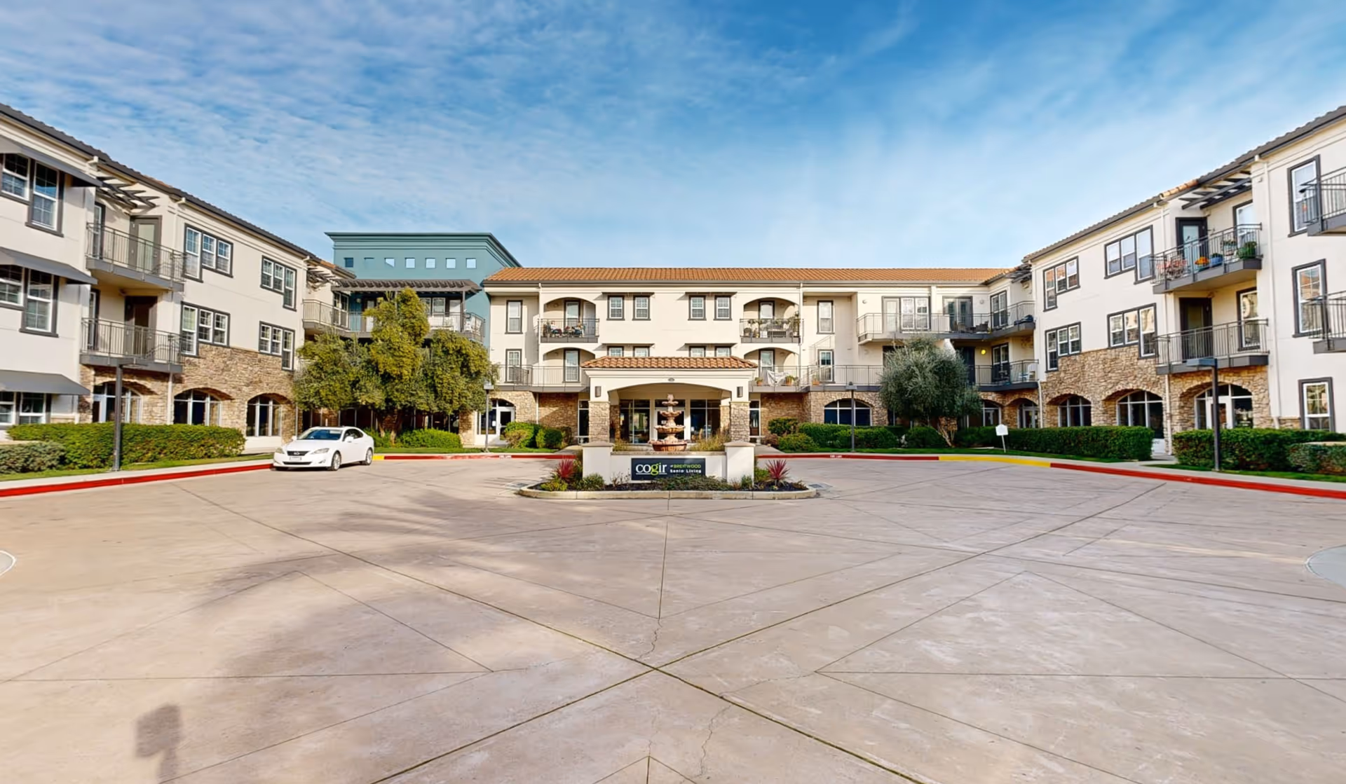 Exterior front view of a multi-story senior living facility with balconies, a central entrance with a covered portico, a fountain, and a sign that reads 'Cogir of Brentwood'. There is a driveway and a parked white car in front of the building, with landscaping including bushes and trees.