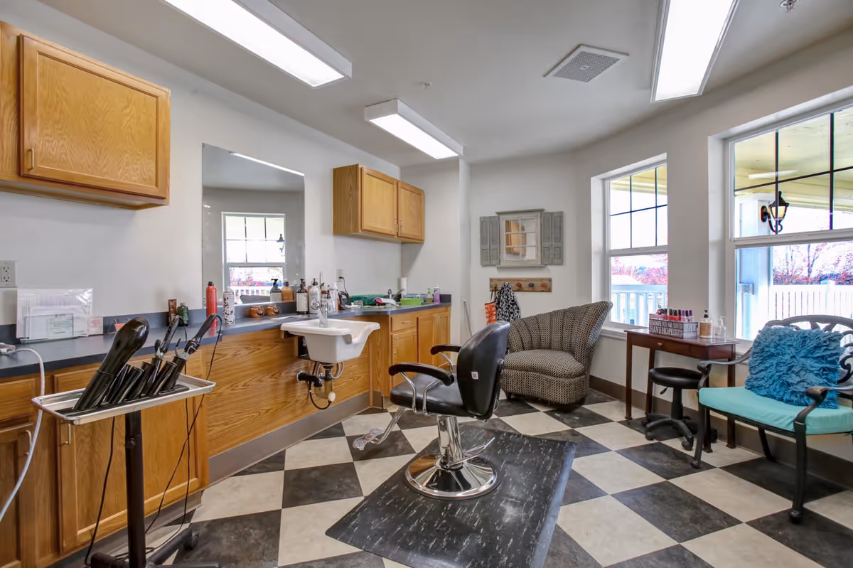 Interior view of a senior living facility hair salon with a black salon chair in front of a sink and mirror, wooden cabinets, hair styling tools on a stand, a patterned armchair, and a seating area with a chair and a small table near large windows.