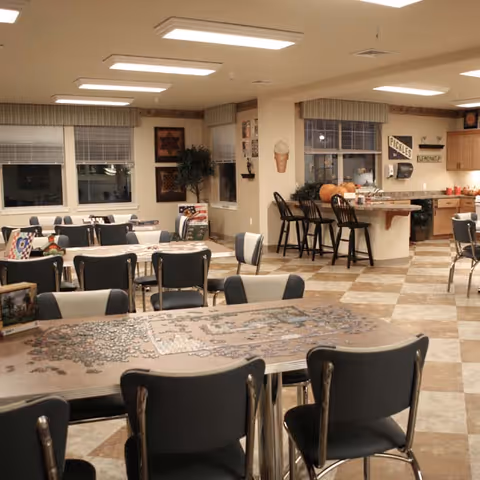 Community dining and activity room with tables and chairs, a jigsaw puzzle on a table, and a kitchen counter with bar stools.