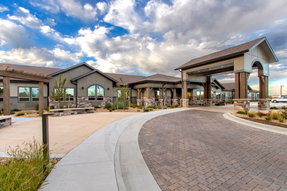 Front exterior view of Aspens at Fort Collins Memory Care facility showing a large covered entrance with stone and wood accents, a curved brick driveway, landscaped areas with small trees and shrubs, and a partly cloudy sky.