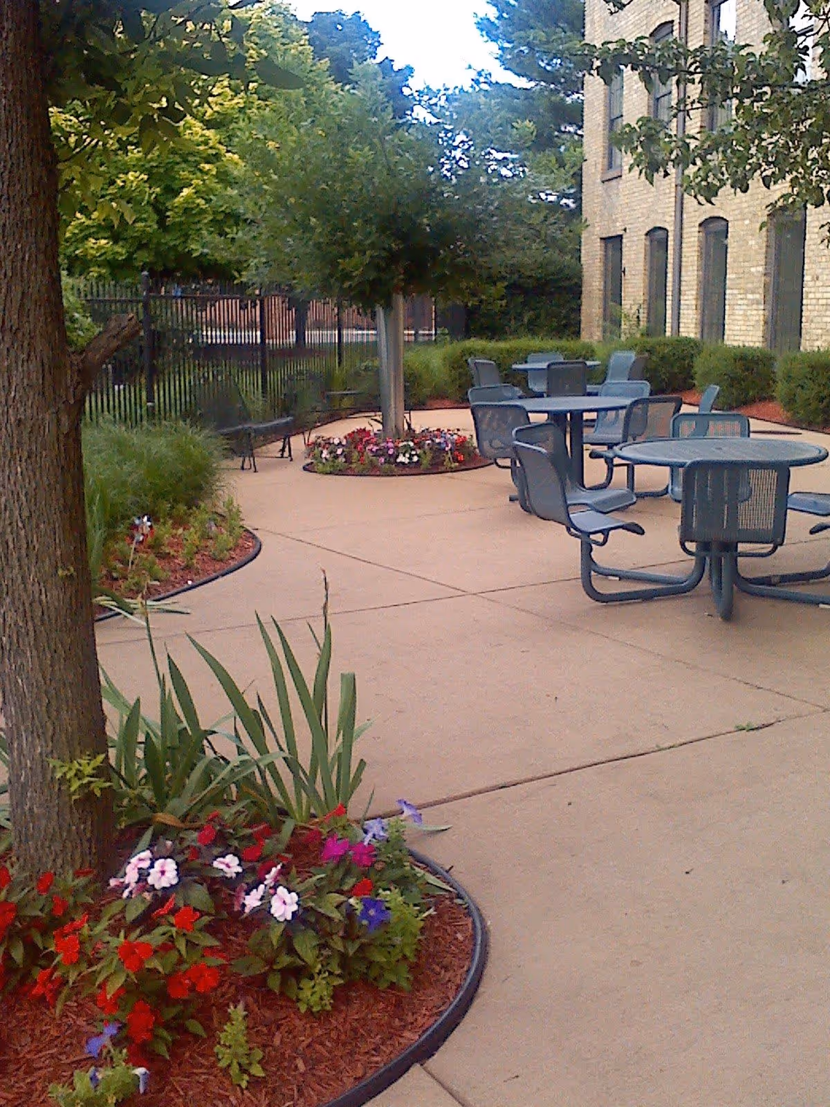 Outdoor patio area with round tables and chairs on a concrete surface, surrounded by landscaped flower beds with colorful flowers and trees. A brick building is visible on the right side, and a black metal fence is in the background.