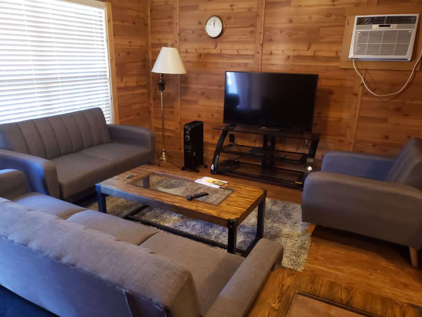 A cozy living room with wooden walls featuring a flat-screen TV on a black stand, a floor lamp, an air conditioner mounted on the wall, and three gray upholstered sofas arranged around a wooden coffee table with a glass inset. A clock is mounted on the wall above the TV, and a gray rug is on the wooden floor.