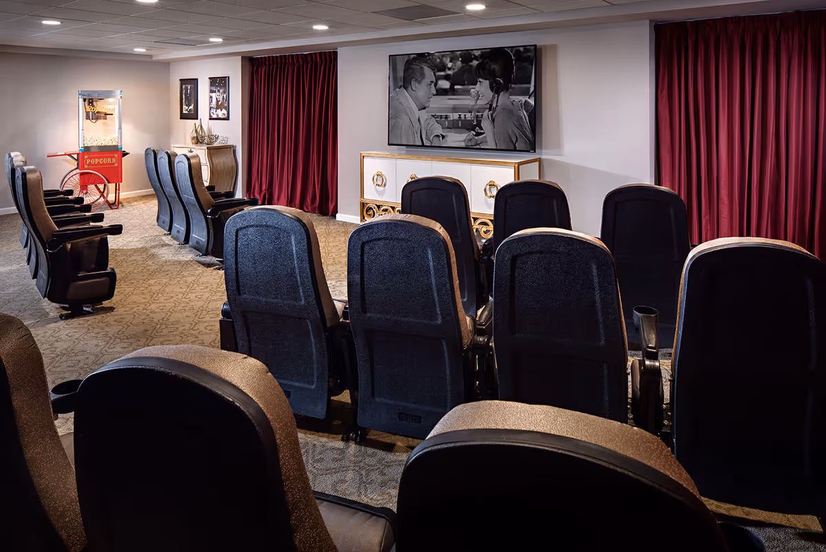 A small movie theater room with several rows of black cushioned chairs facing a large flat-screen TV mounted on the wall. The TV displays a black and white movie scene of a man and woman talking. To the left, there is a red popcorn machine on a stand. The room has carpeted floors, recessed ceiling lights, and red curtains on the walls.