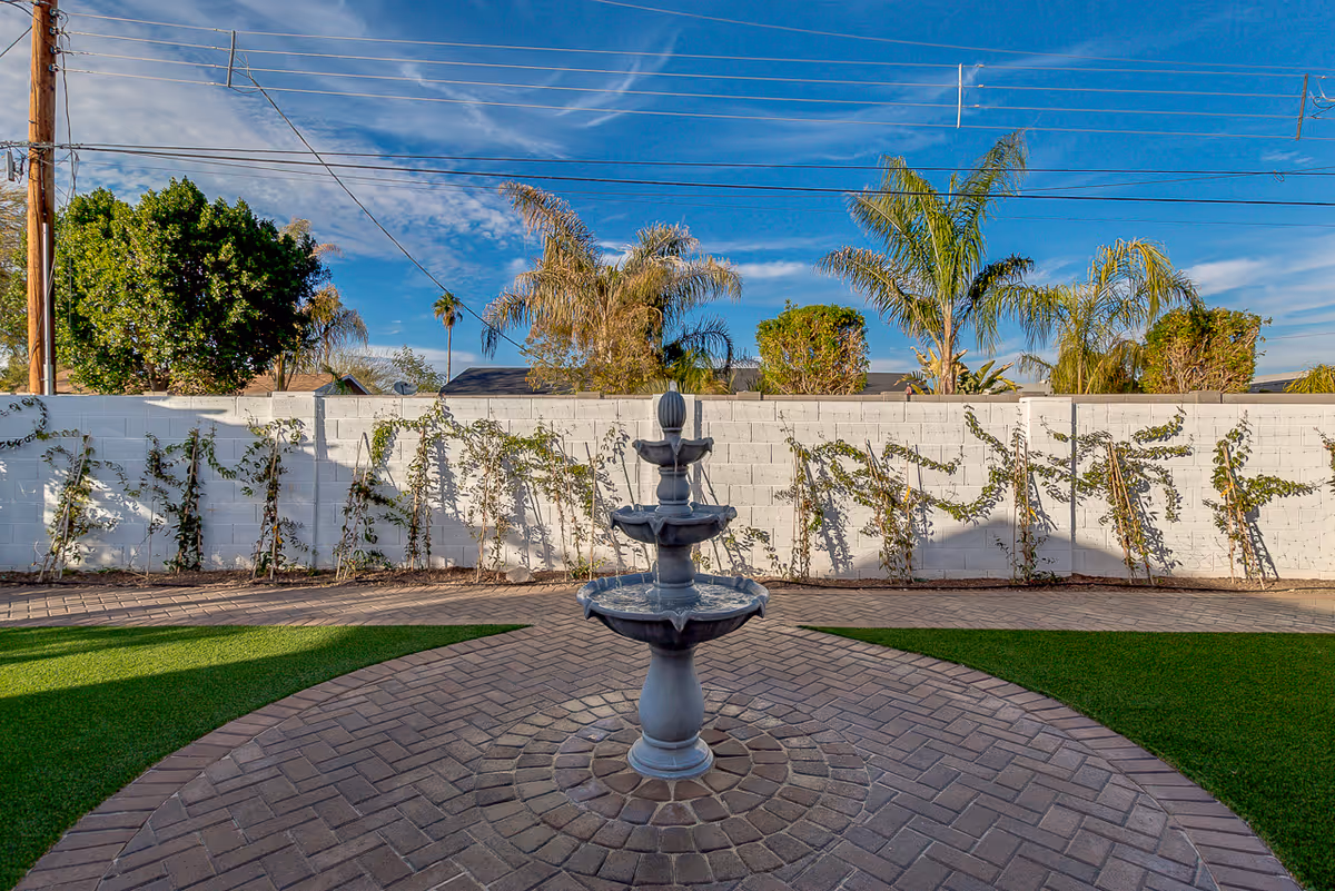 Outdoor courtyard area with a three-tiered stone fountain in the center, surrounded by a circular paved brick pattern and artificial grass. A white brick wall with climbing plants and palm trees is visible in the background under a blue sky with some clouds.