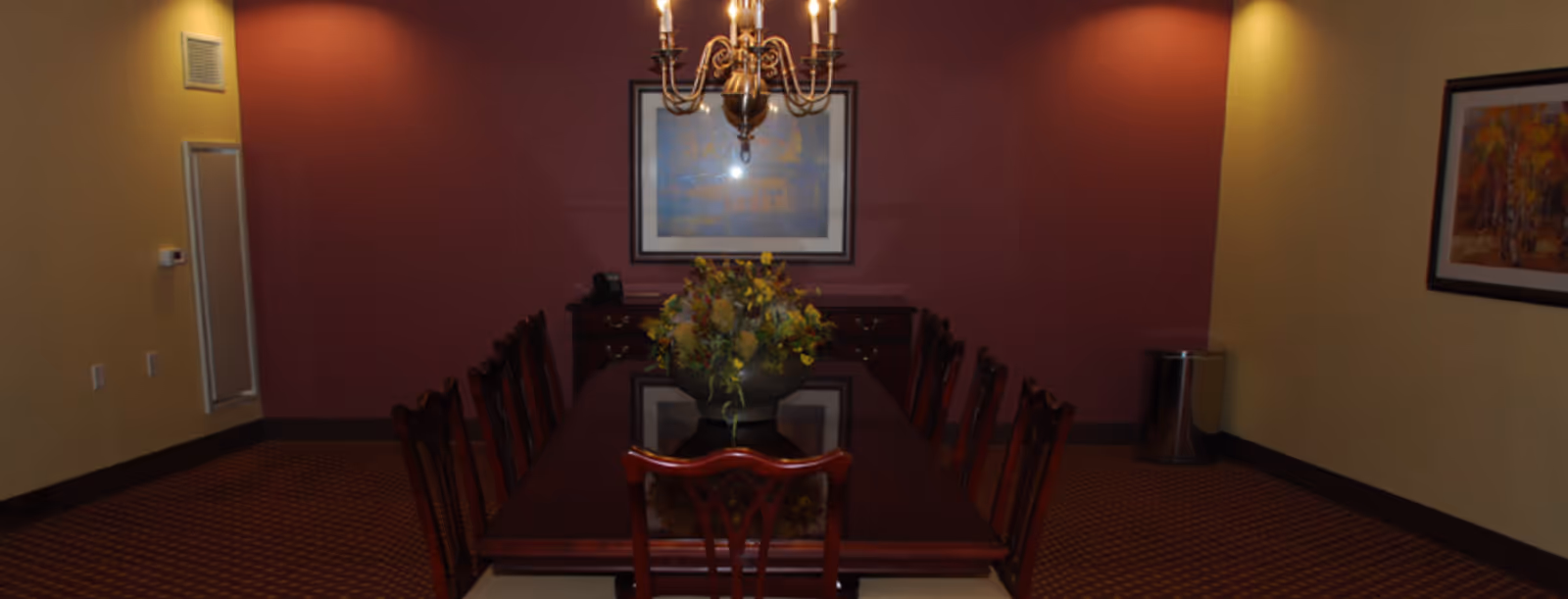 Formal dining room with a long dark wooden table, chairs, chandelier, and a floral centerpiece against maroon and yellow walls.