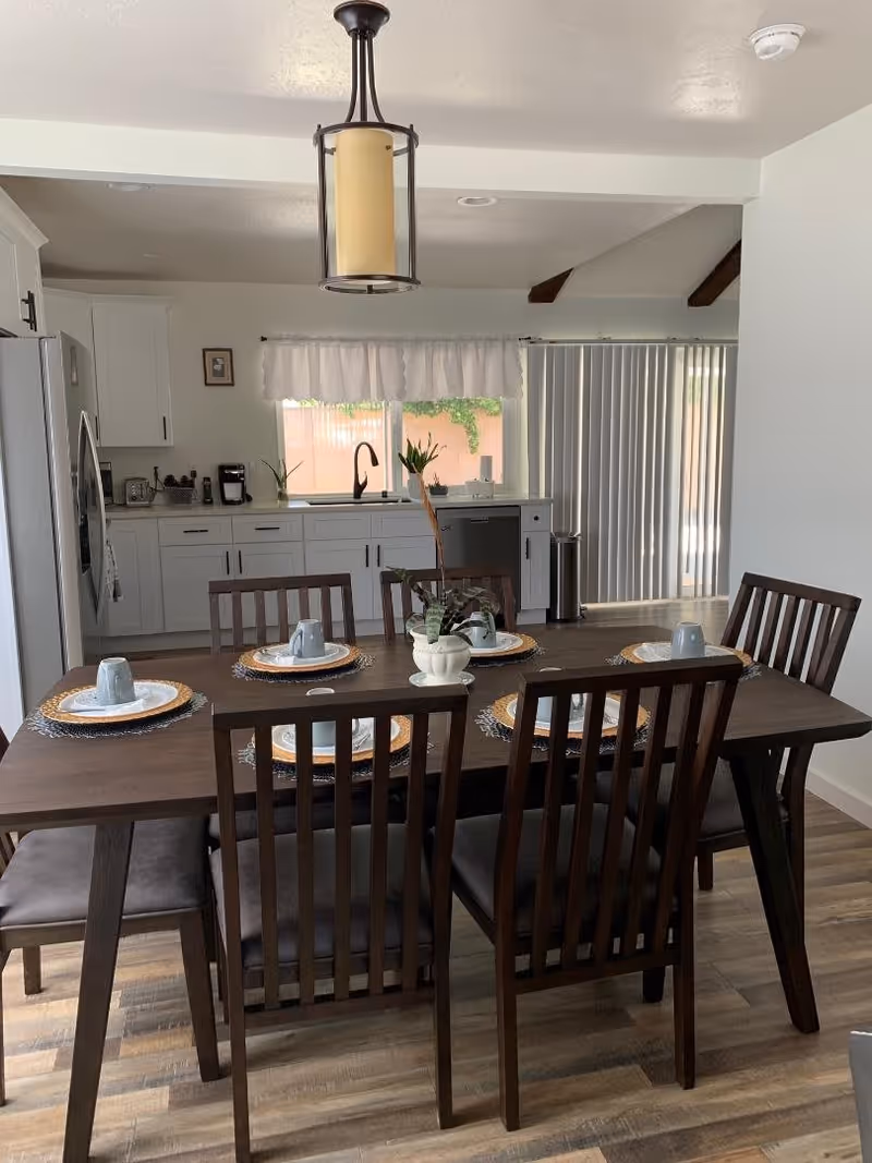 A dining area with a dark wooden table set for six people, each place setting includes a plate, a woven placemat, and a gray cup. The dining table is surrounded by six matching wooden chairs with cushioned seats. In the background, there is a kitchen with white cabinets, a stainless steel refrigerator, a sink under a window with a white valance, and a sliding glass door with vertical blinds. A modern pendant light fixture hangs above the dining table.