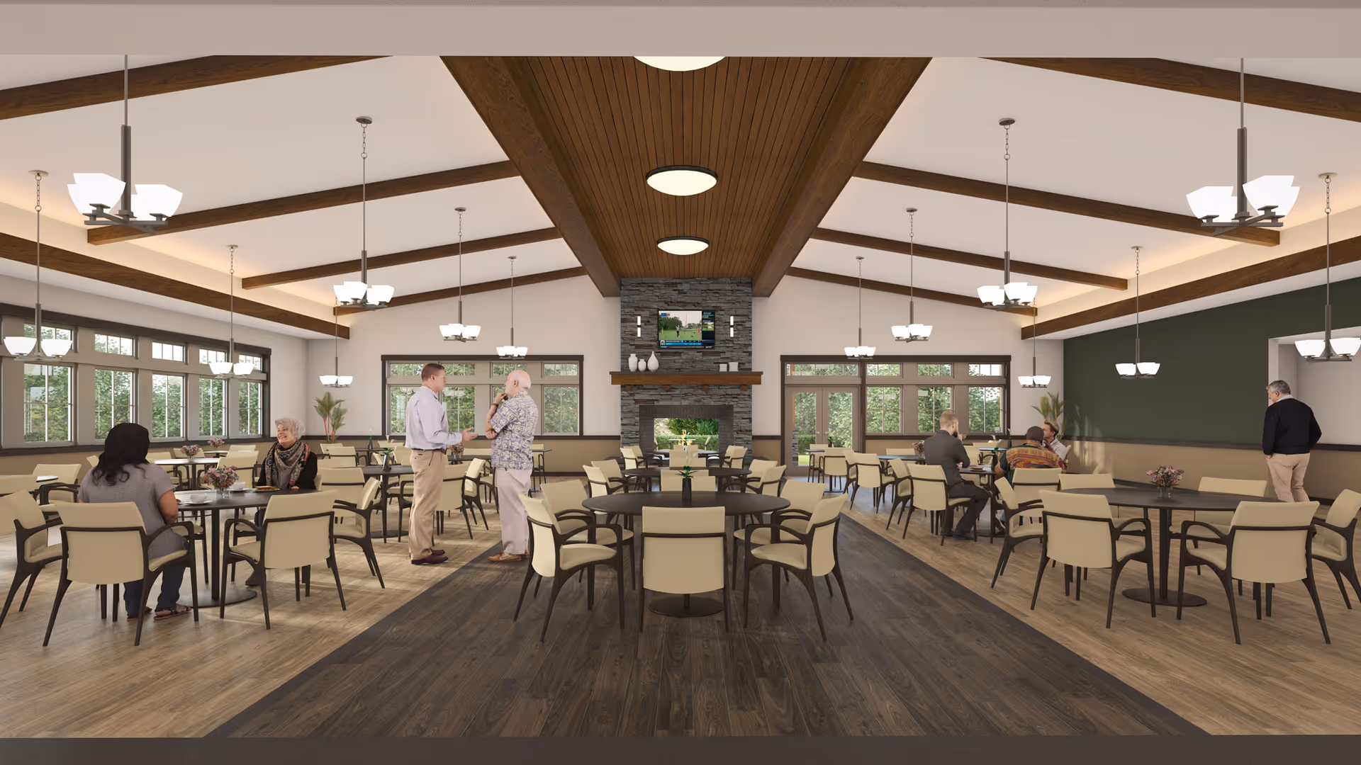 A spacious dining room with multiple round tables and beige chairs. Several people are seated and standing, engaging in conversation. The room features large windows letting in natural light, wooden ceiling beams, and a stone fireplace with a TV mounted above it. Modern pendant lights hang from the ceiling.
