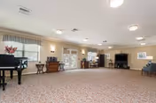 A spacious common area with patterned carpet flooring, beige walls, and ceiling lights. The room features a black grand piano on the left, several wooden tables and chairs, a large flat-screen TV on a stand, and windows with blinds allowing natural light to enter.