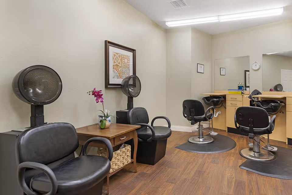 Interior view of a salon area in a senior living facility with two black hair drying chairs on the left, a wooden side table with a purple orchid, and three black salon chairs in front of mirrors and sinks on the right. The floor is wood, and the walls are light-colored with framed artwork and a clock.