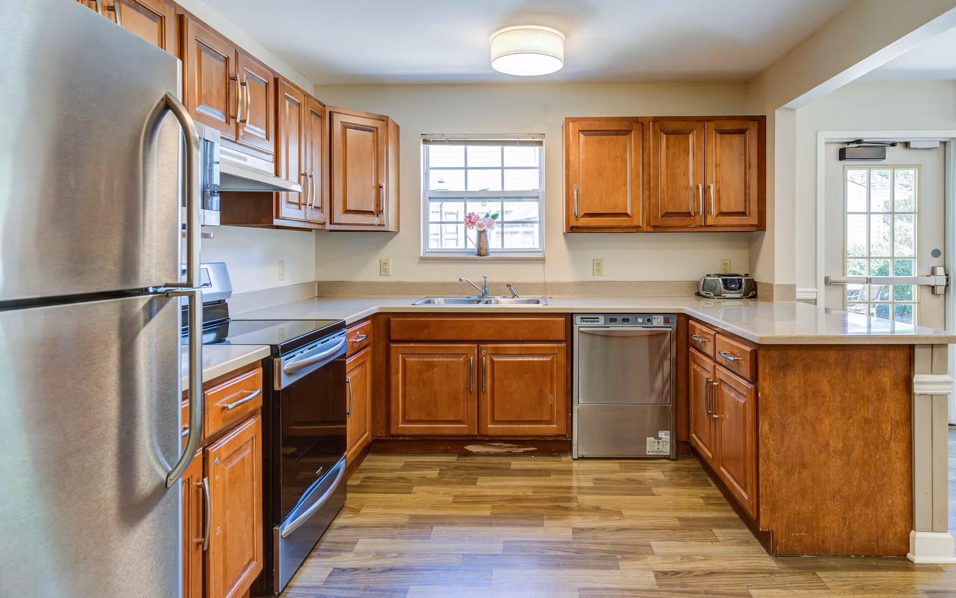 A clean, well-lit kitchen with wooden cabinets, stainless steel refrigerator, oven, and dishwasher. There is a window above the sink with a small vase of flowers on the windowsill. The floor is wood, and there is a door with glass panels leading outside on the right side.