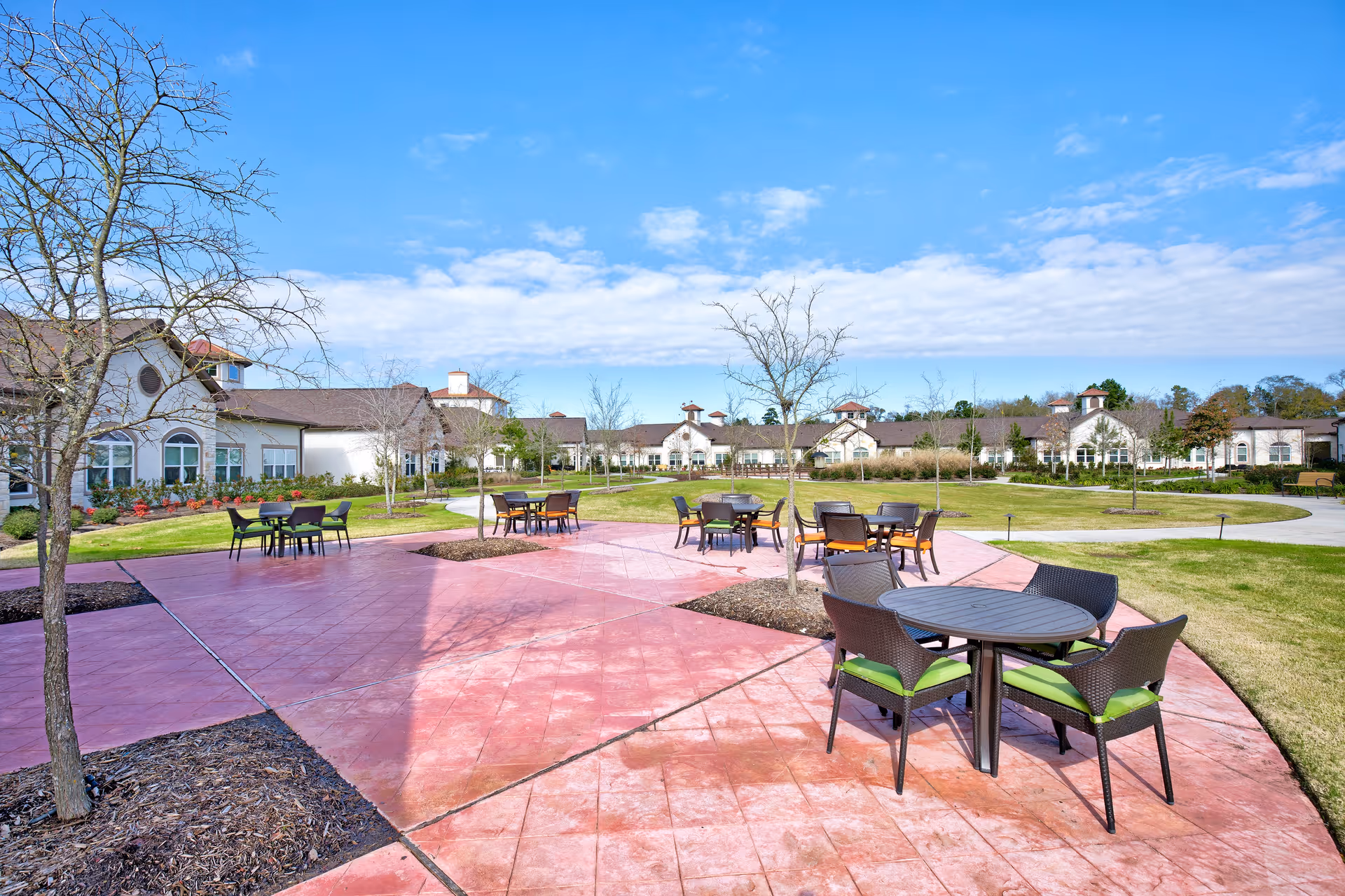 Outdoor patio area at Woodhaven Village with several round tables and chairs arranged on a red paved surface. The patio is surrounded by grass, small trees, and a large building with multiple windows in the background under a partly cloudy blue sky.
