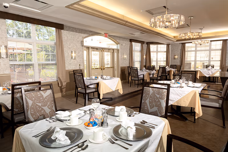 A bright and elegant dining room with multiple tables set for a meal. Each table has white tablecloths with beige underlays, silver plates, white napkins, cups, and cutlery. The room features large windows with blinds and curtains, decorative wall sconces, and modern chandeliers hanging from the ceiling. The entrance door with glass panels is visible in the background.