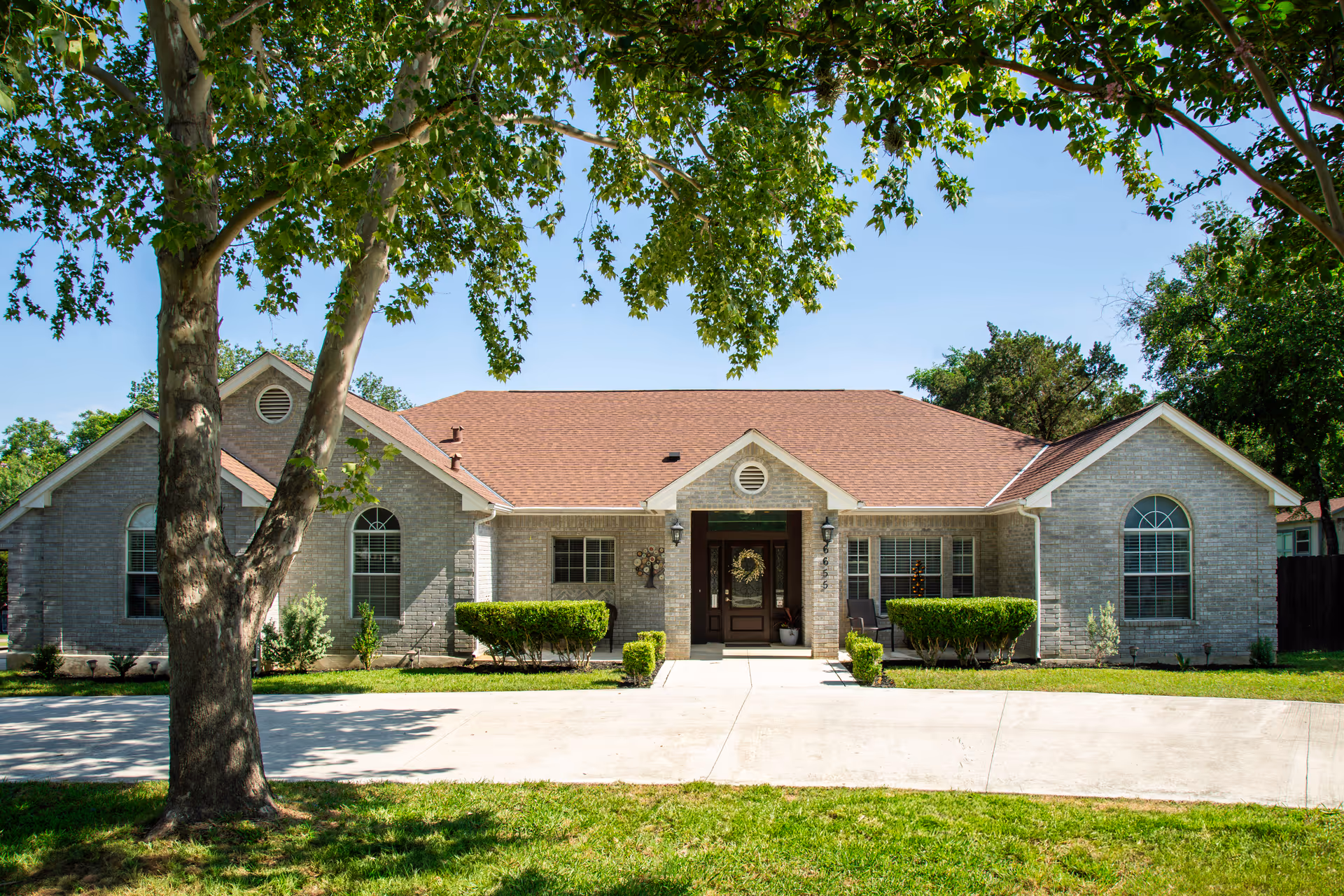 Front exterior view of a single-story brick building with a brown shingled roof, large windows with arched tops, and a central entrance door decorated with a wreath. The building is surrounded by green bushes and trees, with a concrete driveway and a grassy lawn in the foreground under a clear blue sky.