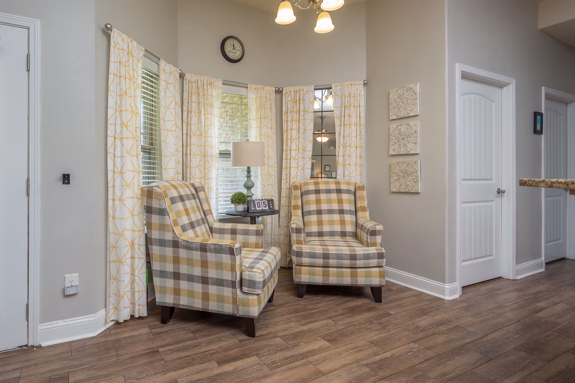 A cozy corner in a living area featuring two plaid upholstered armchairs facing each other with a small round table between them. The table holds a lamp, a small plant, and a flip clock. The space is well-lit with natural light coming through windows covered with white and yellow patterned curtains. The walls are painted light gray and decorated with three square wall art pieces. The floor is wooden, and there are two white doors visible on the right side.