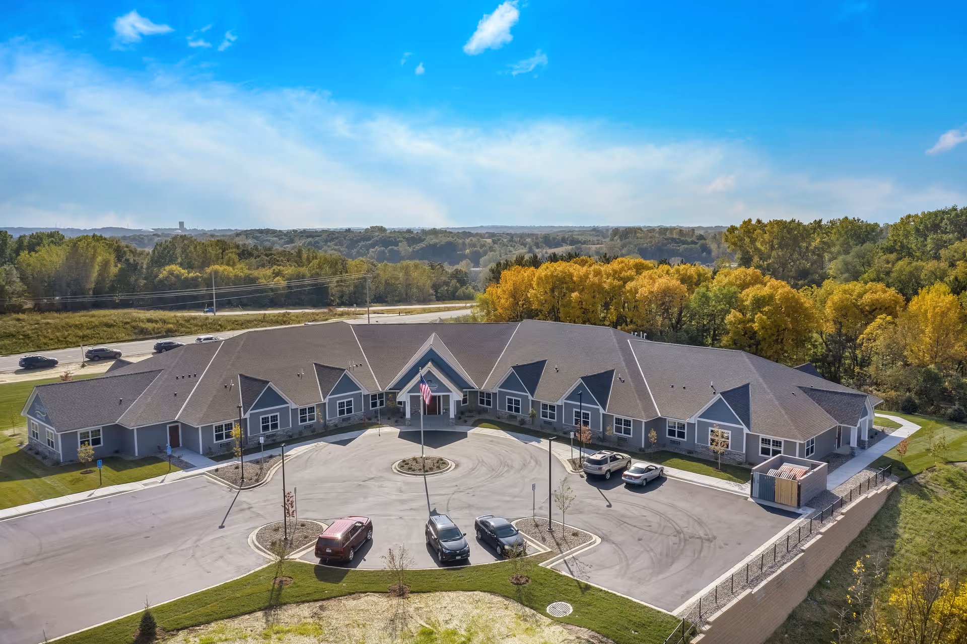 Aerial view of Suite Living Senior Care of Burnsville, a single-story building with a large circular driveway and several parked cars. The building is surrounded by green trees and open land under a blue sky with a few clouds.