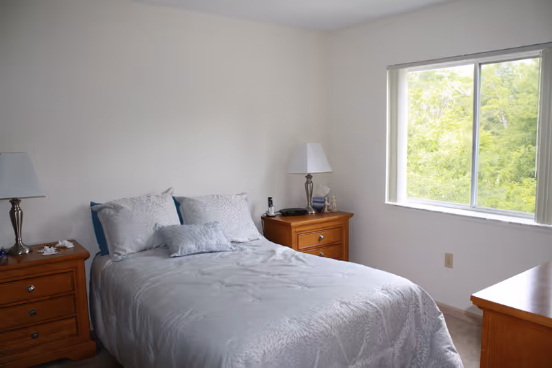 A bright bedroom with a made double bed, matching wooden nightstands, lamps, and a window overlooking trees.