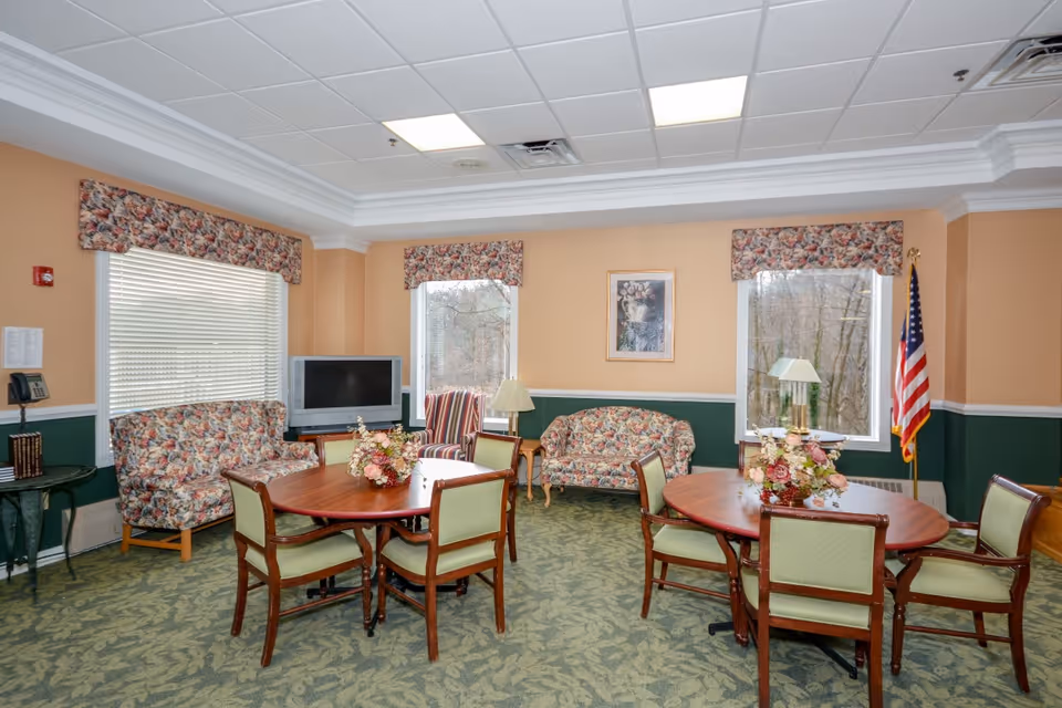 Bright communal sitting room with round wooden tables and chairs, floral-upholstered sofas, a television, and windows.