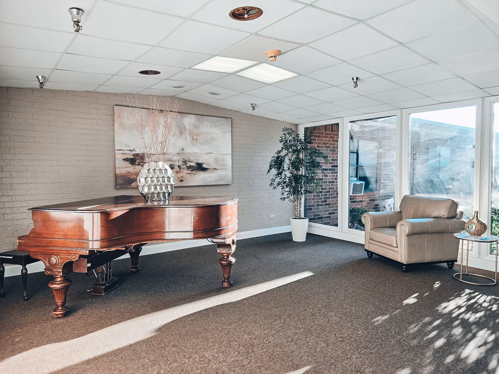 A bright room with large windows letting in natural light, featuring a polished wooden grand piano with a decorative vase on top, a beige armchair next to a small round side table with a decorative item, a potted plant in the corner, and a large abstract painting on the wall.