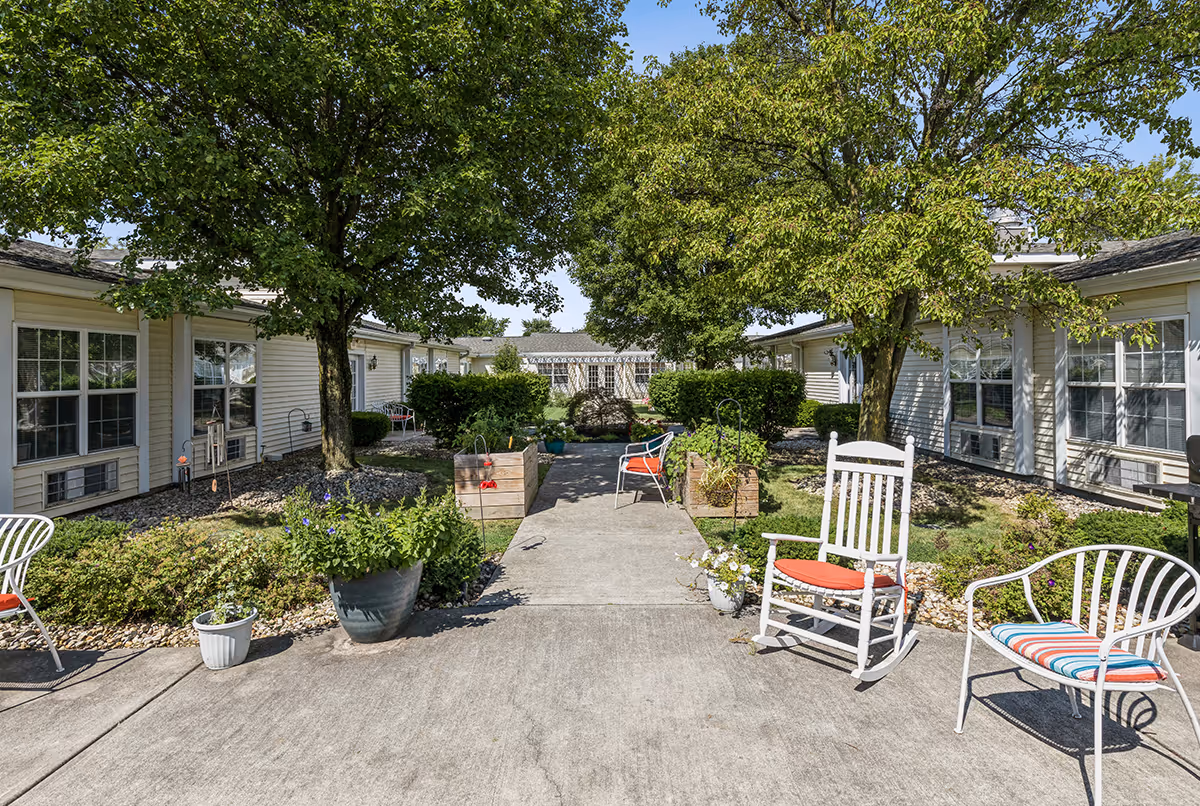 A sunny courtyard with a central paved walkway, rocking chair and patio chairs, potted plants and trees, flanked by single-story residential buildings.