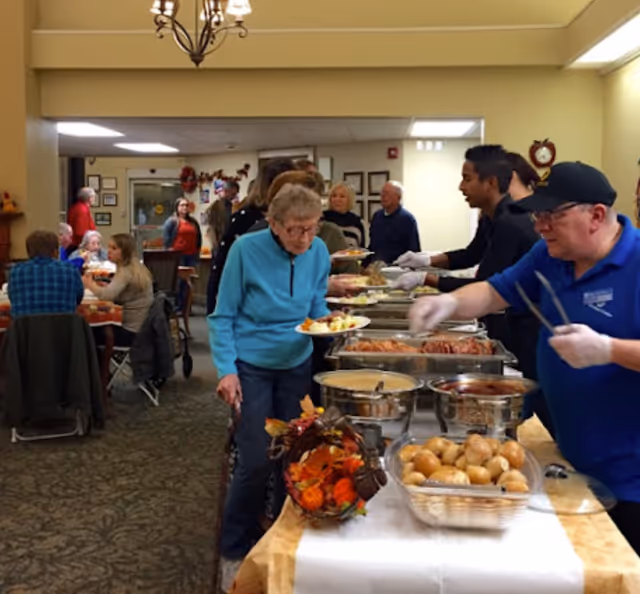 An elderly woman with a cane is serving herself food from a buffet table in a dining area. Several people are seated at tables in the background, and two staff members wearing gloves are assisting with the food service. The room is warmly lit and decorated with autumn-themed decor.