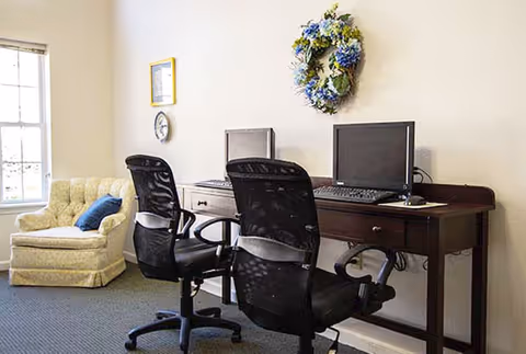A small office area with two black mesh office chairs in front of a dark wooden desk holding two computer monitors and keyboards. To the left, there is a beige upholstered armchair with a blue cushion near a window with blinds. A decorative wreath with blue and white flowers hangs on the wall above the desk.