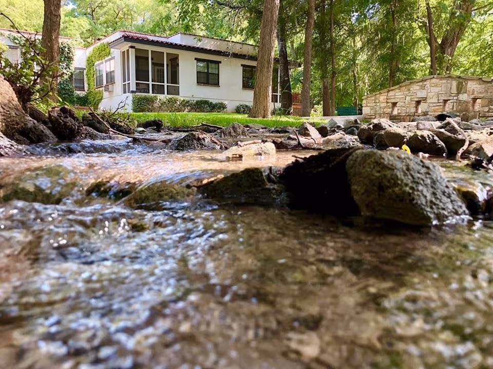 A low-angle view of a small flowing creek with rocks and clear water in the foreground, surrounded by green trees and grass. In the background, there is a white building with a screened porch and a stone structure, all under a canopy of trees.