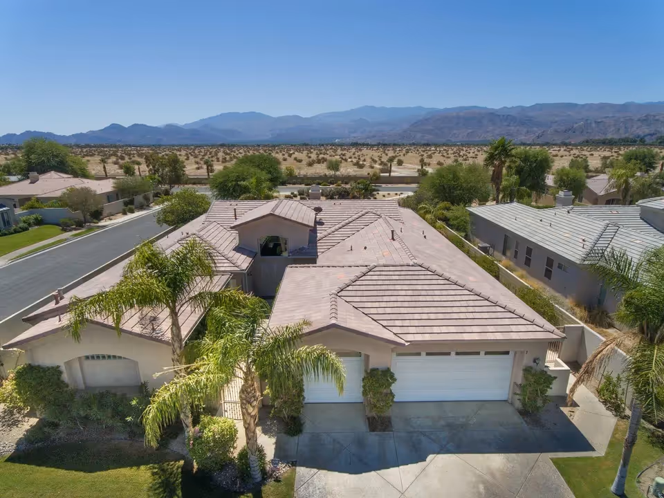 Aerial view of a single-story residential building with a tiled roof, surrounded by palm trees and greenery. The building has a three-car garage and is situated in a desert landscape with mountains in the background under a clear blue sky.