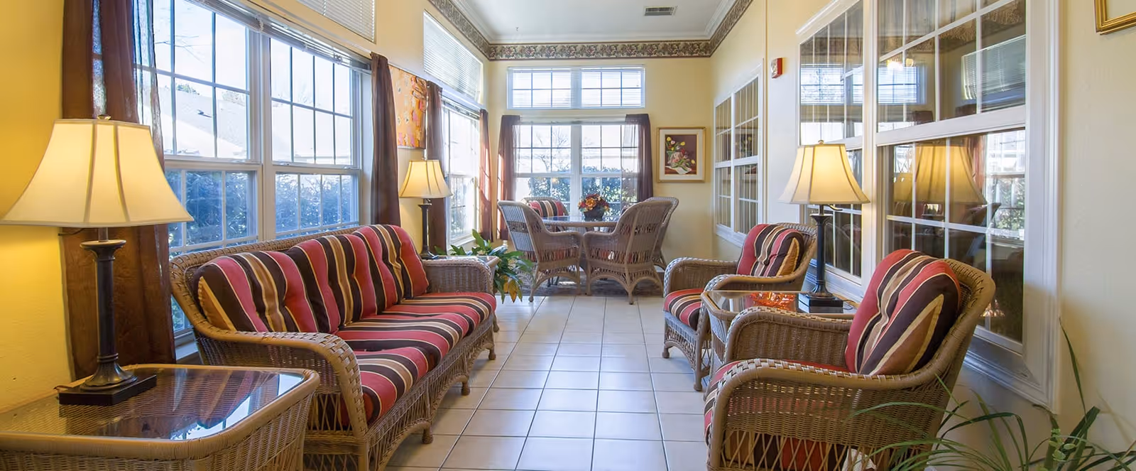 A bright sunroom with large windows on two sides, furnished with wicker furniture including a striped cushioned sofa, two armchairs, and a small table with a lamp. At the far end, there is a wicker dining table with four chairs and a flower arrangement on top. The room has tiled flooring and light-colored walls with framed artwork and several table lamps providing warm lighting.