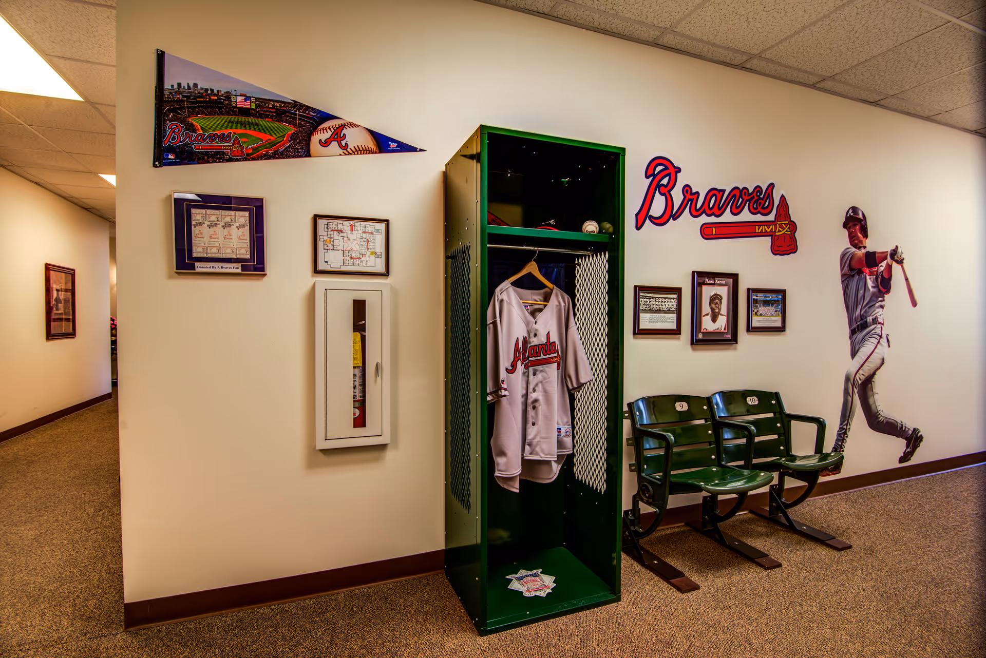 Interior hallway decorated with Atlanta Braves baseball memorabilia including a green locker with a Braves jersey hanging inside, framed pictures, a pennant, a wall decal of a baseball player swinging a bat, and two green stadium seats numbered 9 and 10.