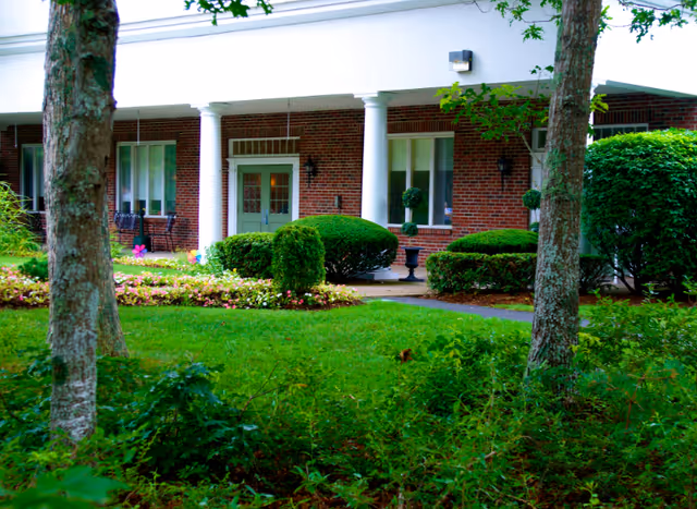 Exterior view of a brick building with white columns and a green door, surrounded by neatly trimmed bushes, flower beds, and trees in a grassy area.