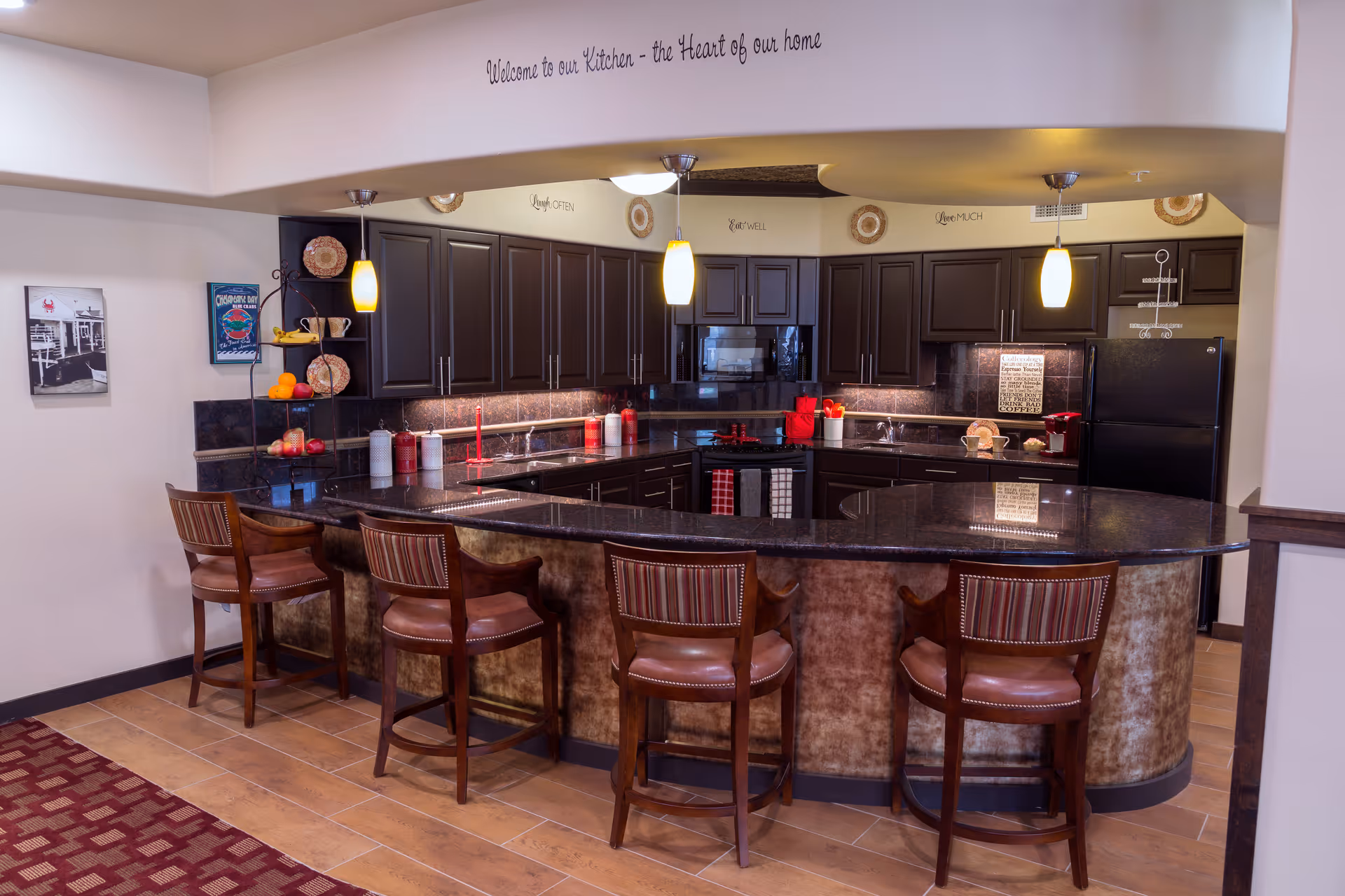 Communal kitchen with a curved granite countertop bar, four wooden barstools, dark cabinets, and pendant lights.
