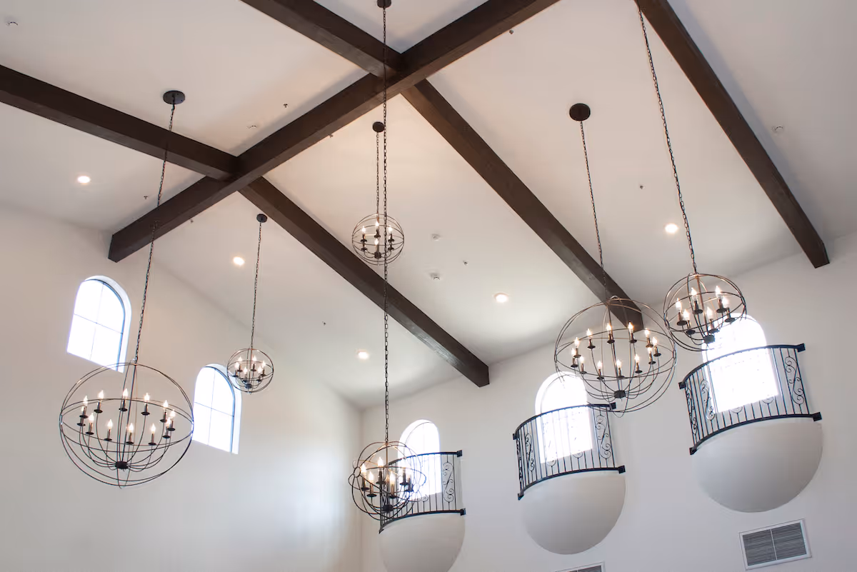 Vaulted interior ceiling with exposed wooden beams, multiple spherical chandeliers, arched windows and decorative iron balcony railings.