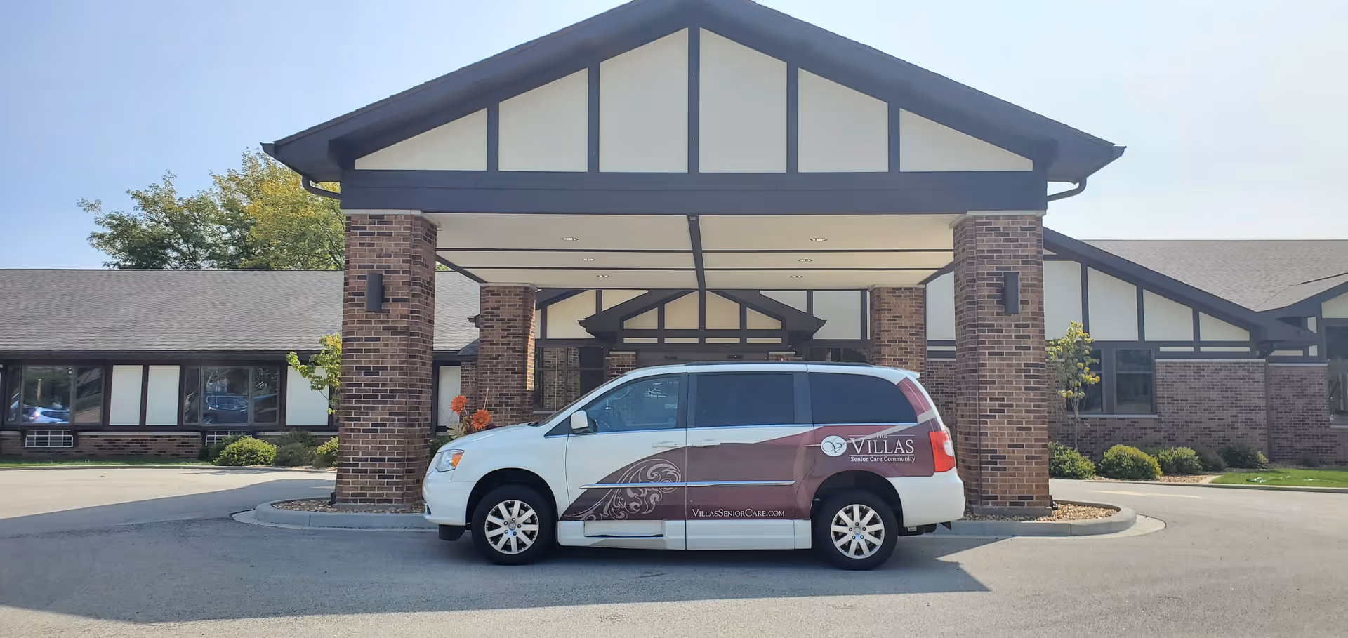 Front exterior view of a senior care community building with a covered entrance supported by brick pillars. A white and maroon van with the logo and text 'Villas Senior Care Community' is parked under the entrance canopy.