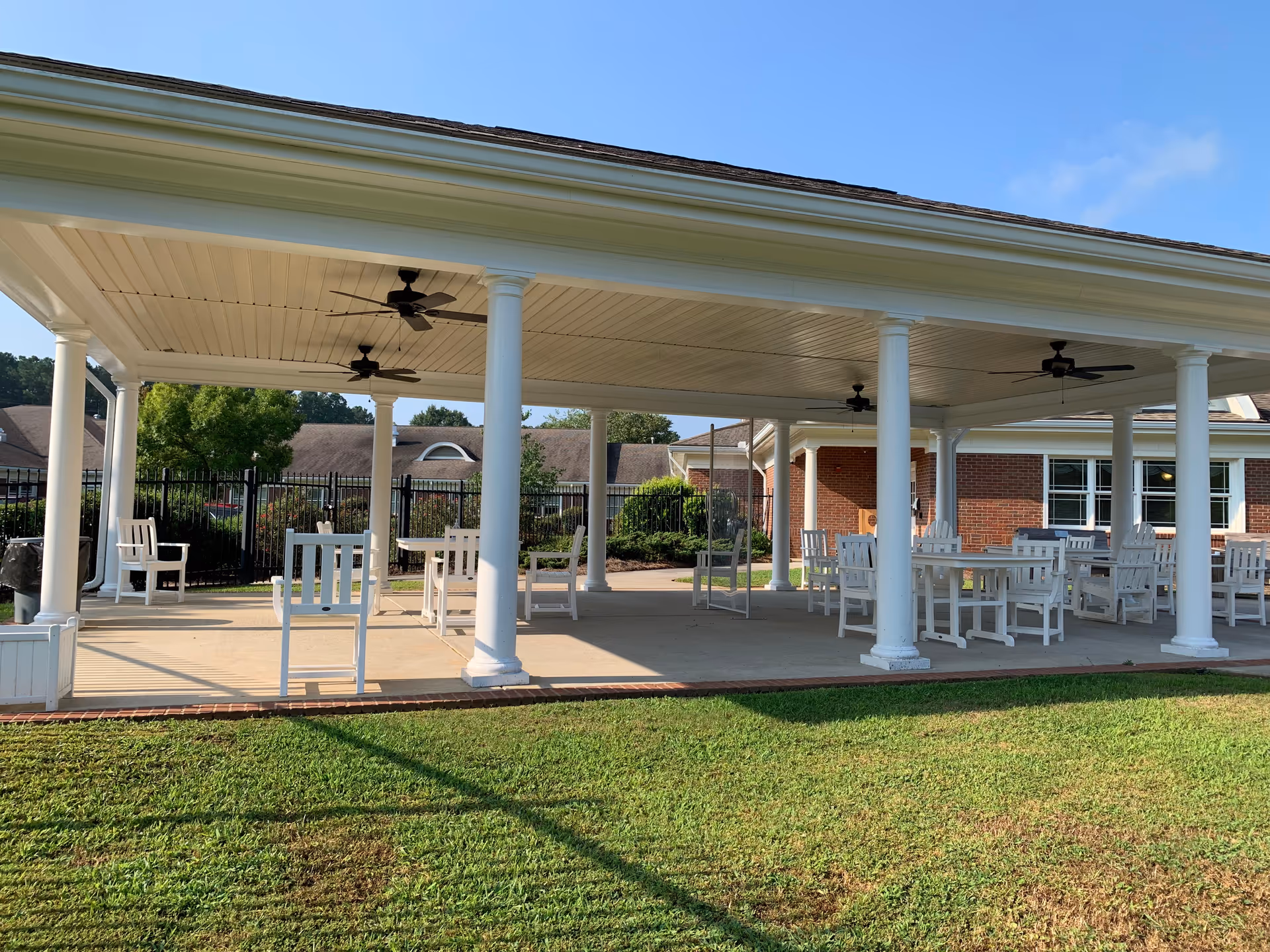 Covered outdoor patio with white columns, ceiling fans, and white chairs and tables on a grassy lawn in front of a brick building.