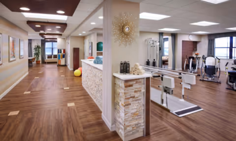 Interior view of a rehab and health care facility showing a hallway with wooden flooring and a reception or check-in counter with towels and water bottles. To the right, there is a physical therapy area with parallel bars and exercise equipment. The space is well-lit with ceiling lights and has large windows allowing natural light.